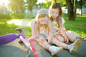 Adorable girl comforting her little sister after she fell off her scooter at summer park. Child getting hurt while riding a kick