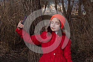Adorable Girl, dressed in red jacket, poses for making selfie or photo of herself with smartphone