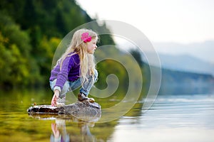Adorable girl playing by Hallstatter See lake in Austria on warm summer day. Cute child having fun splashing water and throwing st