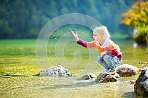 Adorable girl playing by Konigssee lake in Germany on warm summer day. Cute child having fun feeding ducks and throwing stones int