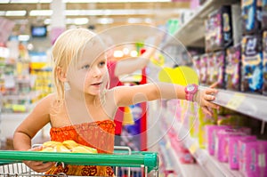 Adorable girl in shopping cart looks at goods on shelves in supermarket