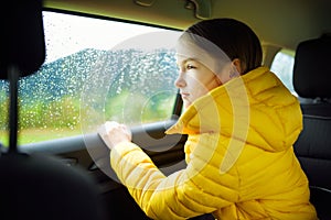 Adorable girl sitting in a car and looking outside on rainy autumn day. Child entertaining herserf on a road trip.