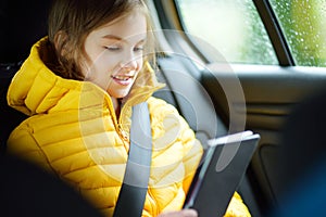 Adorable girl sitting in a car and reading her ebook on rainy autumn day. Child entertaining herserf on a road trip.