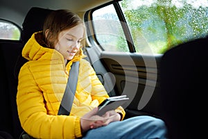 Adorable girl sitting in a car and reading her ebook on rainy autumn day. Child entertaining herserf on a road trip.