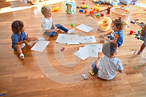 Adorable group of toddlers sitting on the floor drawing using paper and pencil around lots of toys at kindergarten