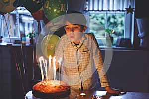 Adorable happy blond little kid boy celebrating his birthday. Child blowing candles on homemade baked cake, indoor