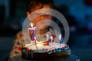 Adorable happy blond little kid boy celebrating his birthday. Child blowing seven candles on homemade baked cake, indoor