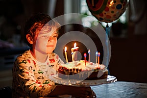 Adorable happy blond little kid boy celebrating his birthday. Child blowing seven candles on homemade baked cake, indoor