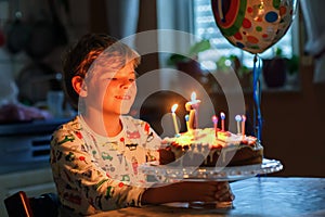 Adorable happy blond little kid boy celebrating his birthday. Child blowing seven candles on homemade baked cake, indoor