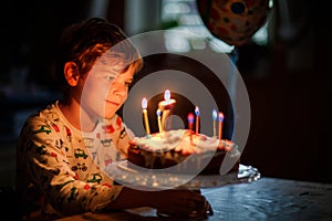 Adorable happy blond little kid boy celebrating his birthday. Child blowing seven candles on homemade baked cake, indoor