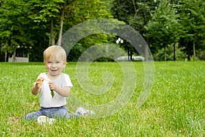 Adorable happy caucasian blonde infant about 1 year old, sitting on grass in park, in summer, playing with leaf