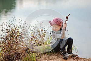 Adorable happy child girl plays with stick on river side in sunny day