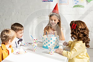 Adorable happy kids sitting at party table with cake while celebrating birthday together.