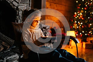 Adorable happy little blonde child girl opening magical Christmas box with gift sitting on rocking chair in xmas night