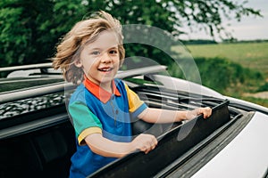 Adorable happy little boy stands in open car sunroof during trip at summer.