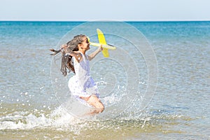 Adorable happy little girl wearing a white dress running through the sea water and playing with the yellow to