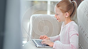 Adorable happy teen girl chatting typing on keyboard using laptop pc sitting sofa