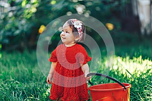 Adorable happy toddler girl with knitten flower crown wearing a red dress enjoying picnic in a beautiful blooming fruit garden