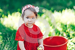 Adorable happy toddler girl with knitten flower crown wearing a red dress enjoying picnic in a beautiful blooming fruit garden