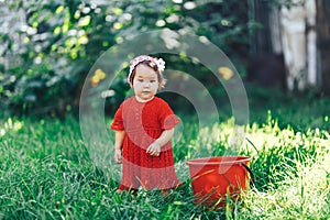 Adorable happy toddler girl with knitten flower crown wearing a red dress enjoying picnic in a beautiful blooming fruit garden