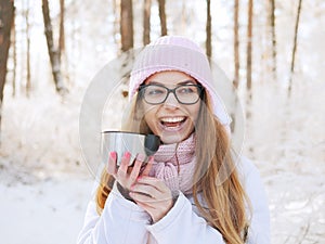 Adorable happy young blonde woman in pink knitted hat scarf having fun drinking hot tea from thermos cup snowy winter park forest