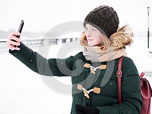 Adorable happy young redhead woman in green parka hat having fun at snowy winter exploring river pier doing photos on smartphone