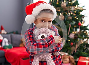 Adorable hispanic boy hugging teddy bear standing by christmas tree at home