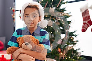 Adorable hispanic boy hugging teddy bear standing by christmas tree at home
