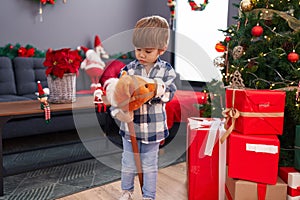 Adorable hispanic boy playing with horse toy standing by christmas tree at home