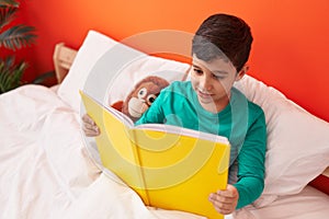 Adorable hispanic boy reading book sitting on bed at bedroom