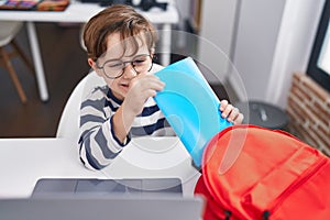 Adorable hispanic boy student holding book of backpack at classroom