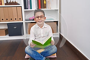 Adorable hispanic boy student smiling confident reading book at library school