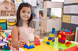 Adorable hispanic girl playing with construction blocks sitting on table at kindergarten