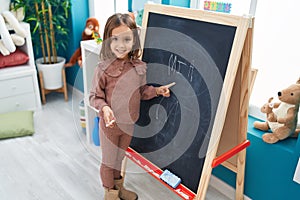 Adorable hispanic girl preschool student smiling confident writing name on blackboard at kindergarten