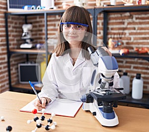 Adorable hispanic girl student using microscope writing on noteboo at laboratory classroom