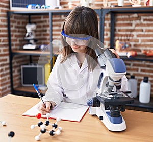 Adorable hispanic girl student using microscope writing on noteboo at laboratory classroom