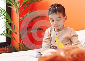 Adorable hispanic toddler reading book sitting on bed at bedroom