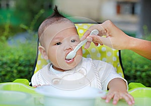 Adorable infant baby boy sitting in the chair and eating food for the first time. Hand of mother feeding food into baby mouth by