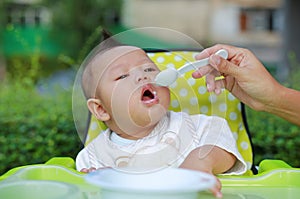 Adorable infant baby boy sitting in the chair and eating food for the first time. Hand of mother feeding food into baby mouth by
