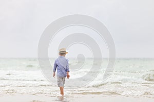 Adorable kid boy in straw hat and sun glasses walking on ocean beach and playing with waves. Vacations by the sea
