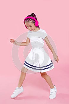 Adorable kid dancing with headphones, over pink background. Happy ittle girl dancing in studio. Full length portrait.