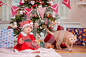 An adorable kid dressed as Santa Claus plays near a decorated Christmas tree in the living room. A cat walks around the gifts