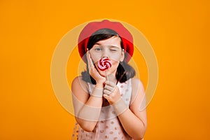 Adorable kid eating candy with pleasure. Studio shot of brunette little girl with lollipop isolated on yellow background