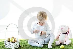 Kid holding white box while sitting on green grass near toy rabbit and straw basket with Easter eggs isolated on white