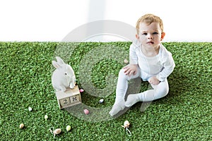 Kid sitting on green grass near wooden calendar with 28 April date, decorative rabbit and colorful Easter eggs isolated