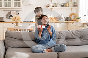 Adorable kid son greeting mom with mother day, birthday with gift box closing eyes of excited mum