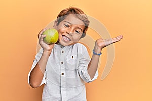 Adorable latin kid holding green apple celebrating achievement with happy smile and winner expression with raised hand