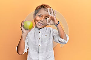 Adorable latin kid holding green apple doing ok sign with fingers, smiling friendly gesturing excellent symbol