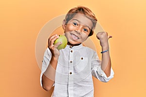 Adorable latin kid holding green apple pointing thumb up to the side smiling happy with open mouth