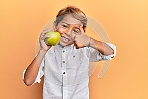 Adorable latin kid holding green apple smiling happy and positive, thumb up doing excellent and approval sign